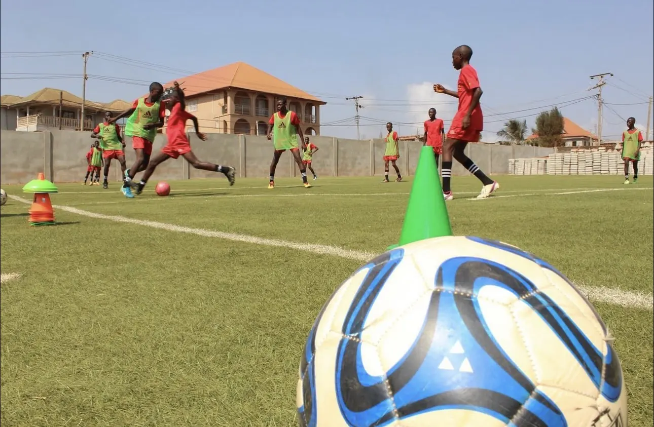 Loma Academy players in red training kit and green bibs doing a dribbling drill on the artificial pitch in Calaba Town